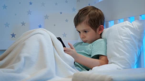 Portret małego skoncentrowanego chłopca leżącego pod kocem w łóżku i korzystającego z tabletu. Dzieci ze smartfonami i gadżetami w nocy — Wideo stockowe