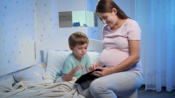 Fiatal mosolygós terhes anya ül mellette a gyermek fekszik az ágyban, és videót néz tabletta számítógépen — Stock videók