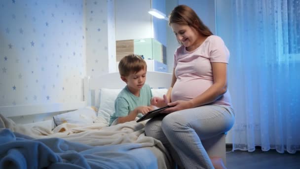 Piccolo bambino ragazzo con giovane madre incinta giocare e guardare cartoni animati su tablet computer a letto prima di andare a dormire — Video Stock