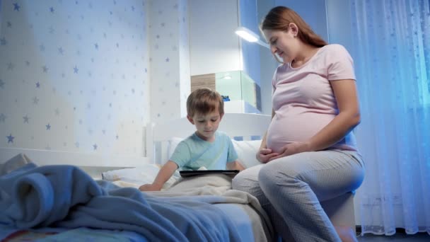 Mujer embarazada joven sentada en la cama de su hijo pequeño y mirándolo jugar juegos en la computadora de la tableta. Niños y adultos usando teléfonos inteligentes y aparatos por la noche durante la hora de acostarse — Vídeos de Stock