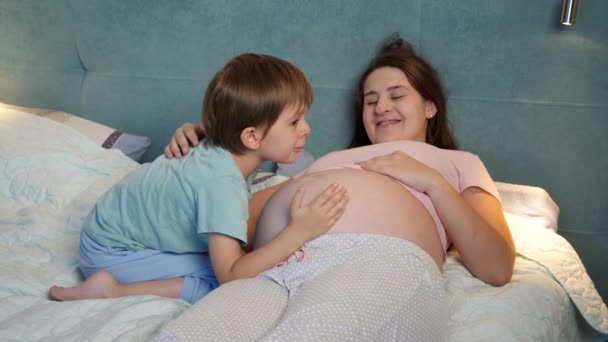 Sonriente niño pequeño en pijama hablando y abrazando al bebé nonato en el vientre grande de la madre embarazada acostada en la cama por la noche — Vídeo de stock
