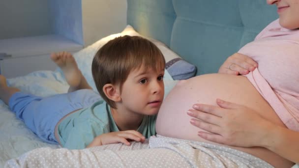 Usmívající se malý chlapec a těhotná matka v pyžamu leží v posteli v noci a mluví, Rodiče mají čas se svými dětmi doma — Stock video