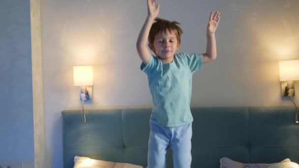 Забавный маленький мальчик в пижаме прыгает и танцует на кровати родителей по ночам. Бессонный и активный мальчик перед сном — стоковое видео