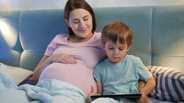 Портрет беременной молодой матери, обнимающей своего маленького сына, лежащего в постели и играющей на планшетном компьютере перед сном. — стоковое видео