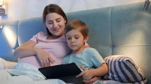 Familia acostada en la cama y viendo dibujos animados en la tableta por la noche. Niño pequeño y madre embarazada usando tableta digital en la cama por la noche — Vídeo de stock