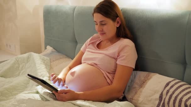 Portret van een lachende zwangere vrouw in pyjama 's die' s nachts in bed liggen en surfen op internet of sociale media op tabletcomputer — Stockvideo