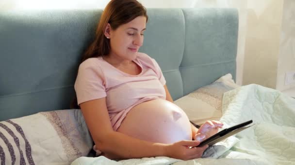 Lächelnde Schwangere im Schlafanzug mit Tablet-Computer und tippender Nachricht, während sie nachts im Bett liegt — Stockvideo