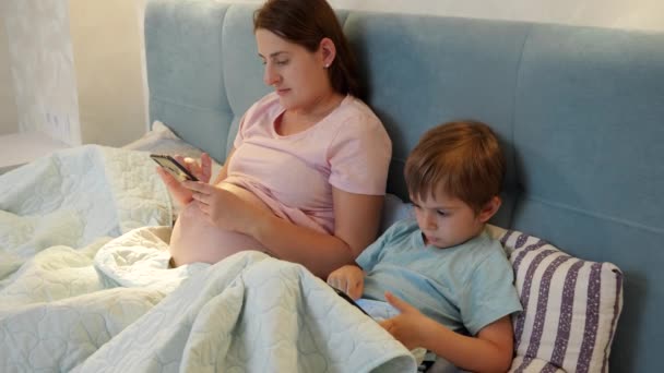 Маленький мальчик с беременной матерью в пижаме просматривает интернет и играет в игры на смартфоне и планшетном компьютере, лежа ночью под одеялом — стоковое видео