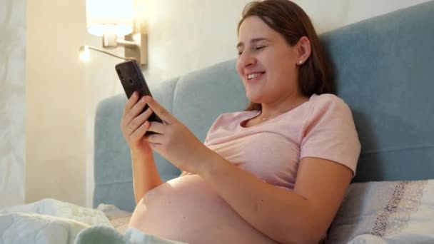Portrét usměvavé těhotné ženy v pyžamu ležící v posteli a mající videokonferenci na smartphonu. Mladá těhotná dívka mluví prostřednictvím videohovoru na telefonu — Stock video