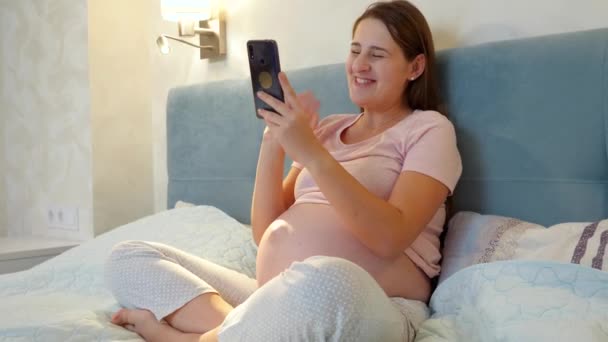 Молодая беременная женщина разговаривает по видеосвязи, сидя ночью на кровати. Беременная девушка разговаривает с другом или врачом через видеоконференцию на смартфоне — стоковое видео