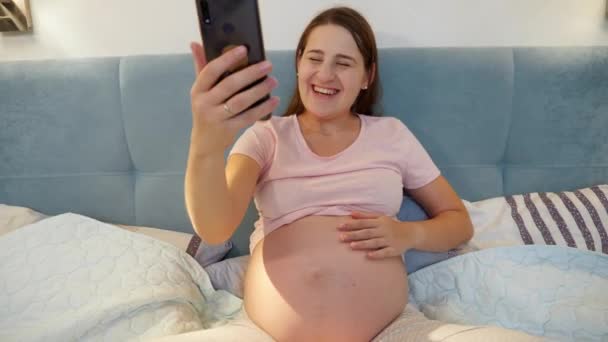 Femme enceinte souriante attendant bébé assis au lit et appelant un ami ou un médecin en vidéoconférence. Femme enceinte utilisant un smartphone — Video