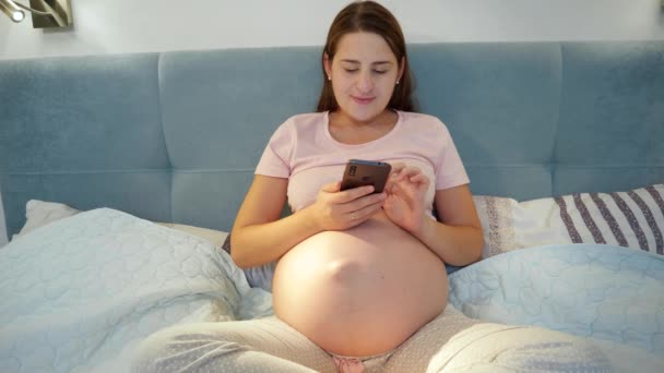 Unge gravide kvinder i pyjamas liggende i sengen og ved hjælp af smartphone. Gravid kvinde med telefon liggende i sengen om natten – Stock-video