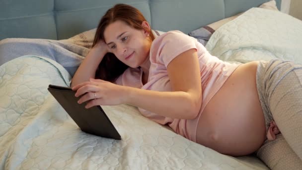 Usmívání těhotné ženy v pyžamu ležící v posteli a prohlížení internetu nebo sociálních médií na tabletu počítače v noci — Stock video