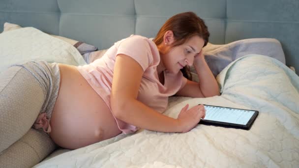 漂亮的孕妇穿着睡衣，在睡前阅读短信，在平板电脑上浏览社交媒体 — 图库视频影像