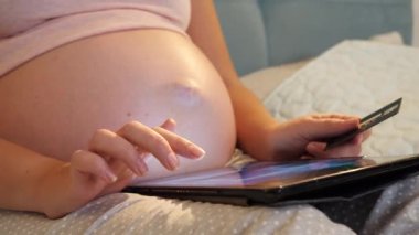 Hamile bir kadının gece yatağında yatıp tablet bilgisayardan online alışveriş yapmasına yakın çekim. Çevrimiçi alışveriş kavramı ve aygıtlar üzerinde e-mağaza