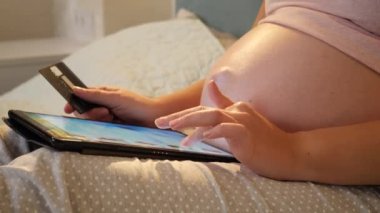 Koca göbekli bir bebeği bekleyen ve tablet bilgisayardan online alışveriş yapan bir kadının yakınlaşması. Çevrimiçi alışveriş kavramı ve aygıtlar üzerinde e-mağaza