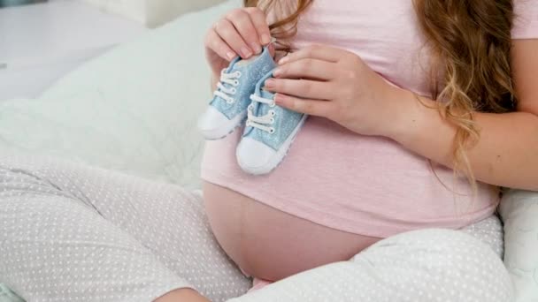 Zbliżenie młodej kobiety w ciąży trzymającej małe dziecięce buty dla niemowląt i chodzącej po dużym brzuchu. Pojęcie ciąży i spodziewania się dziecka — Wideo stockowe
