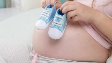 Hamile bir kadını yatakta yatarken ve yeni doğmuş mavi bebek botlarını koca göbeğinde tutarken. Hamilelik kavramı, bebek hazırlamak ve doğurmak