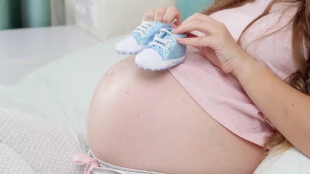 Großaufnahme einer schwangeren Frau, die im Bett liegt und mit Babystiefeln auf großem Bauch tanzt. Konzept von Schwangerschaft und Schwangerschaft — Stockvideo