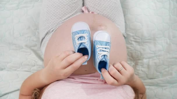 Вид зверху на вагітну жінку з великим животом, що сидить на ліжку і грає з маленькими новонародженими дитячими черевиками. Концепція вагітності, підготовка і очікування дитини — стокове відео