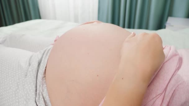 Close-up de jovem grávida deitada na cama no quarto do hospital e acariciando sua barriga grande antes de dar à luz. Conceito de gravidez, preparando e esperando criança. — Vídeo de Stock