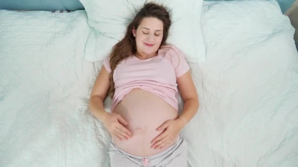 Gyönyörű terhes nő pizsamában pihen az ágyon és simogatja a nagy hasát. Fogalom a terhesség, előkészítése és várandós gyermek otthon — Stock videók