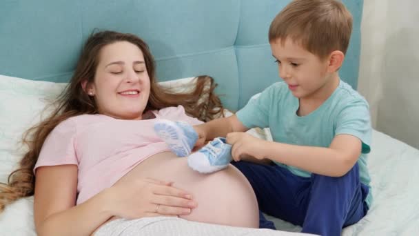 Maličký batole chlapec hrát s modrými botami na velké břicho mladé matky ležící na posteli. Koncepce těhotenství, příprava a očekávání dítěte. — Stock video