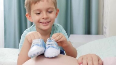 Küçük bebek botlarıyla oynayan mutlu bir çocuğun portresi ve evde yatan hamile bir anne. Hamilelik kavramı, çocuk hazırlama ve hamile kalma.