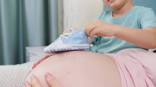 Primer plano del hijo mayor caminando con botas de bebé acroos gran vientre de la madre embarazada acostada en la cama en el dormitorio — Vídeos de Stock