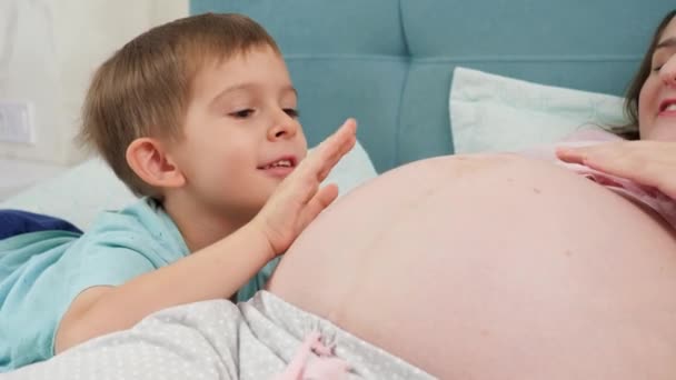 Portrét usměvavého chlapečka hladícího a dotýkajícího se velkého břicha těhotné matky ležící doma na posteli. Koncepce těhotenství, příprava a očekávání dítěte. — Stock video