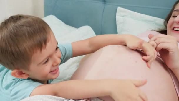 Funny laughing boy playing and tickling young pregnant mother with big belly lying on bed — Stock Video