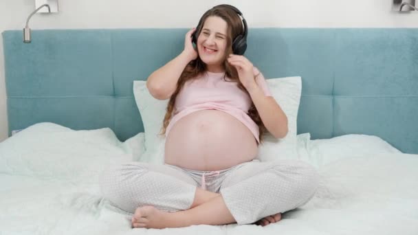 Šťastná usměvavá těhotná žena poslouchá hudbu ve sluchátkách a dává si je na velké břicho. Nenarozené dítě poslouchá klasickou hudbu, zatímco je v matčině břiše. Koncept raného umění a — Stock video