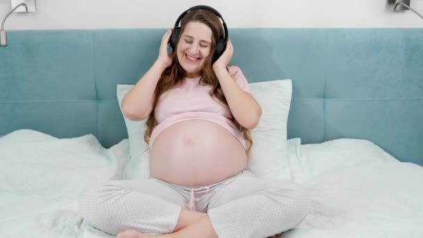 Portret van een lachende zwangere vrouw die geniet van het luisteren naar muziek met een koptelefoon op haar grote buik terwijl ze thuis op bed zit.Ongeboren baby die naar klassieke muziek luistert terwijl ze in moeders buik zit — Stockvideo