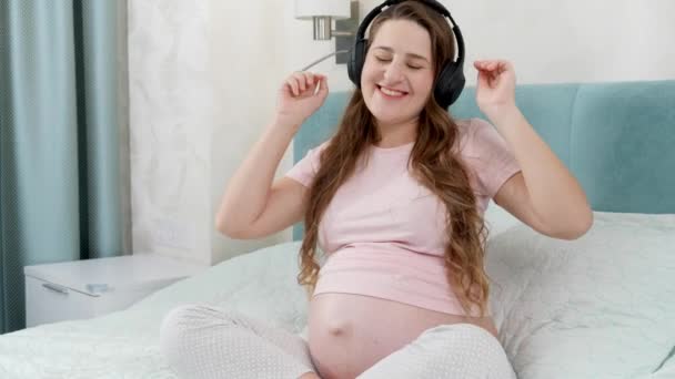 Happy smiling pregnant woman with big belly enjoys listening music in headphones. Unborn baby listening to classic music while being in mothers abdomen. Concept of early art and creativity development — Stock Video