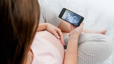 Hamile kadının koca göbeğini okşayıp akıllı telefondan ultrason görüntüsüne baktığı en iyi görüntü. Bebek, hamilelik ve sağlık hizmetleri kavramı.