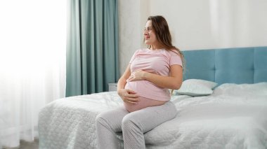 Güzel, gülümseyen, pijamalı hamile bir kadın. Yatakta oturuyor ve koca göbeğini okşuyor.