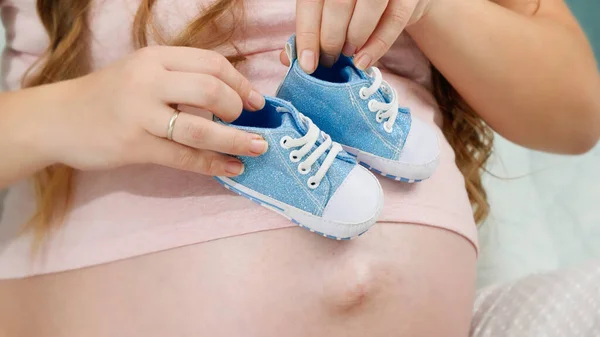Primo piano di bella donna incinta con grande pancia con scarpe per bambini piccoli seduti sul letto. Concetto di gravidanza e attesa del bambino — Foto Stock