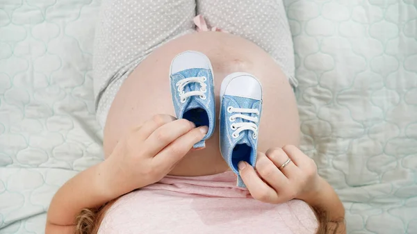 Horní pohled na těhotnou ženu s velkým břichem sedí na posteli a hraje si s malými novorozeneckými botičkami. Koncepce těhotenství, příprava a očekávání dítěte — Stock fotografie