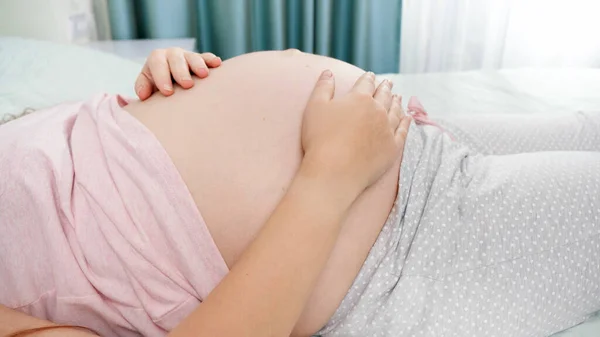 Close-up de jovem grávida deitada na cama no quarto do hospital e acariciando sua barriga grande antes de dar à luz. Conceito de gravidez, preparando e esperando criança. — Fotografia de Stock