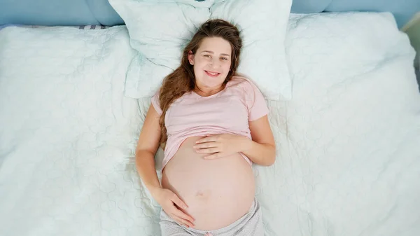 Vacker gravid kvinna i pyjamas vilar på sängen och smeker hennes stora mage. Begreppet graviditet, förbereda och förvänta barn hemma — Stockfoto