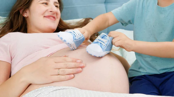 Piccolo bambino ragazzo che gioca con stivali blu bambino sulla grande pancia della giovane madre sdraiata sul letto. Concetto di gravidanza, preparazione e attesa del bambino. — Foto Stock