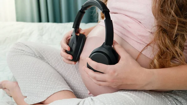 Крупним планом вагітна жінка сидить на ліжку і кладе навушники на її великий живіт. Ненароджена дитина слухає класичну музику під час перебування у матерів живота. Концепція розвитку раннього мистецтва та творчості — стокове фото