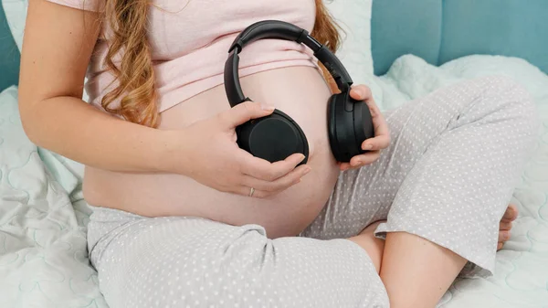 Pijamalı hamile bir kadın müzik dinliyor ve koca göbeğine kulaklık takıyor. Doğmamış bebek anne karnındayken klasik müzik dinliyor. Erken dönem sanat ve yaratıcılık kavramı — Stok fotoğraf