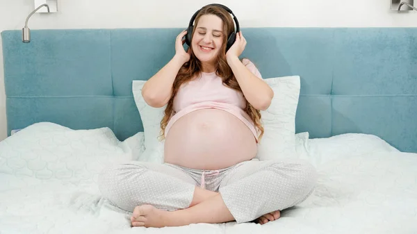 Happy smiling pregnant woman listening to music in headphones and putting them on her big belly. Unborn baby listening to classic music while being in mothers abdomen. Concept of early art and — Stock Photo, Image