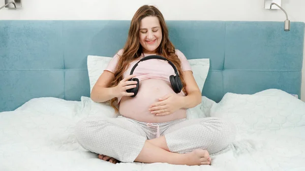 Retrato de mulher grávida sorridente gostando de ouvir música colocando fones de ouvido em sua barriga grande enquanto se senta na cama em casa.Bebê não nascido ouvindo música clássica enquanto está no abdômen das mães — Fotografia de Stock