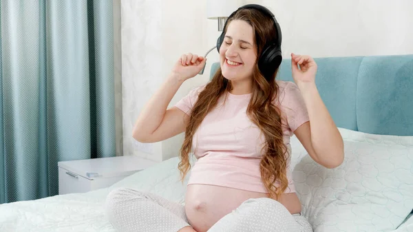 Smiling pregnant woman dancing and enjoying listening music. She puts headphones on her big belly. — Stock Photo, Image