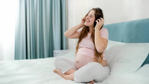 幸せな笑顔妊娠中の女性でパジャマで音楽を聴くとともにヘッドフォンでベッドの上で朝 — ストック写真