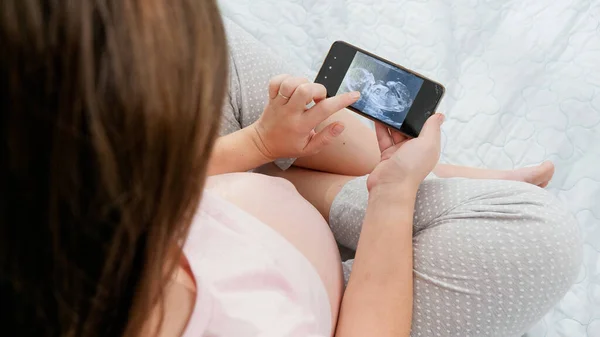 임신 한 여성이 아직 태어나지 않은 아기의 초음파 영상을 보고 큰 배를 쓰다듬는 모습을 지켜보고 있습니다. 출산, 임신 및 건강 관리에 대한 개념. — 스톡 사진