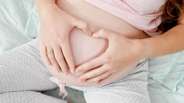 Großaufnahme einer schwangeren Frau, die auf dem Bett sitzt und mit den Händen auf ihrem dicken Bauch Herzform anlegt. Konzept von Kindererziehung, Liebe und Elternschaft. — Stockfoto