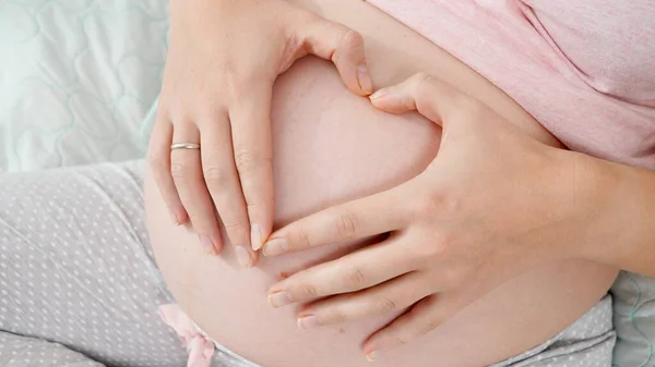 Zbliżenie kobiet w ciąży ręce tworząc kształt serca na dużym brzuchu. Pojęcie spodziewania się dziecka, miłości i rodzicielstwa. — Zdjęcie stockowe
